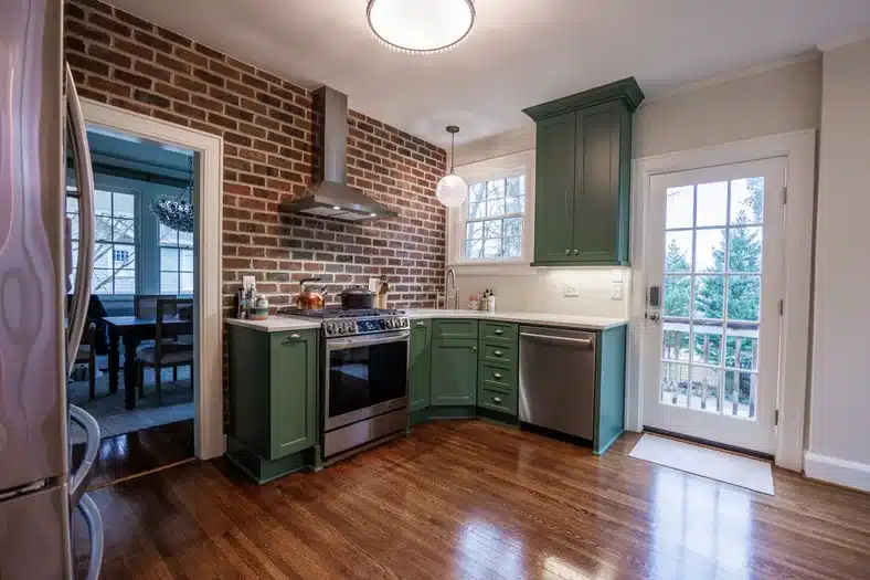 Kitchen with green cabinets, exposed brick wall, stainless steel appliances, and hardwood flooring.