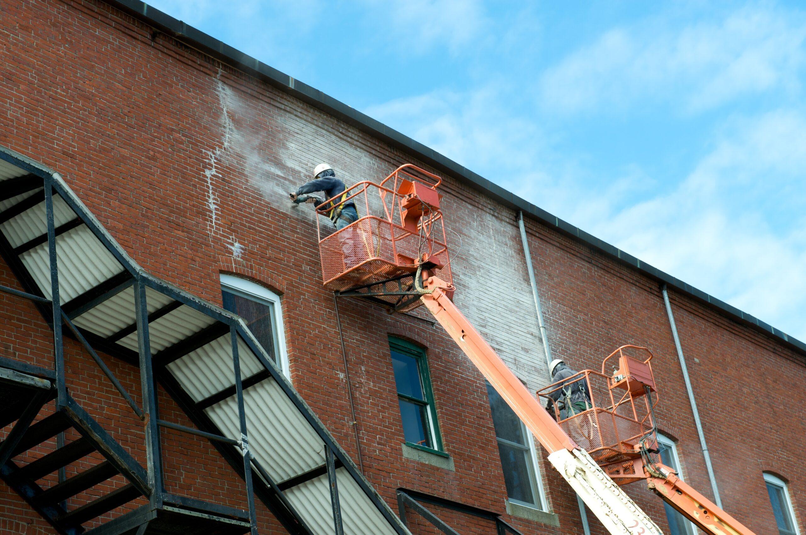 Workers on lift platforms repairing and pressure washing the brick exterior of a commercial building.